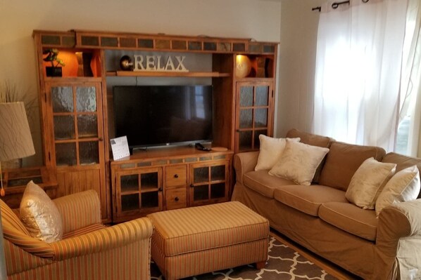 Una sala de estar con una unidad de entretenimiento de madera, un televisor, un sofá marrón, un reposapiés a rayas y un sillón a rayas.