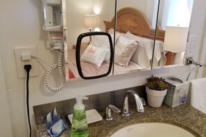 Bagno d'albergo con specchio, lavandino, dispenser per sapone, porta-spazzolino e una pianta in vaso.