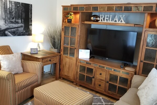 Una sala de estar acogedora con un televisor de pantalla plana grande, armarios de madera, sofás a rayas y un cartel que dice 'RELAX'.