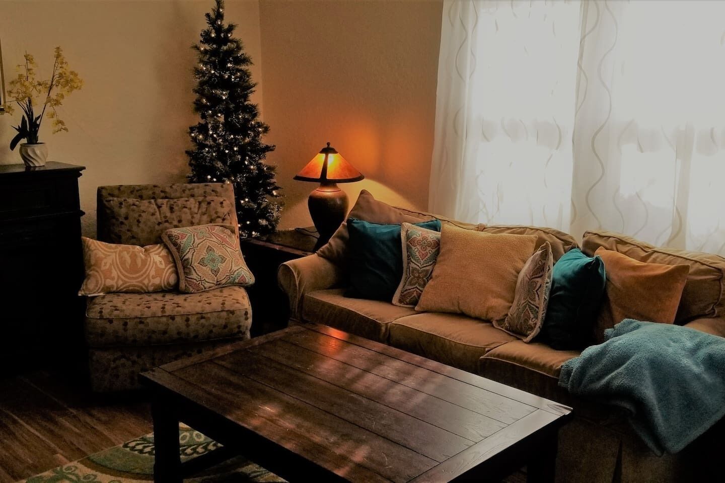 Una sala de estar acogedora con una mesa de centro de madera, un sillón estampado, un sofá con muchos cojines, un árbol de Navidad y una lámpara de pie.