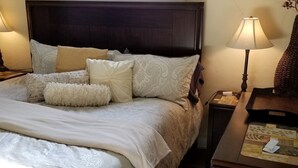 Ein ordentlich bezogenes Bett mit einem gemusterten Überwurf und mehreren Kissen, ein dunkles Kopfteil sowie ein Nachttisch mit Lampe und dekorativer Vase.