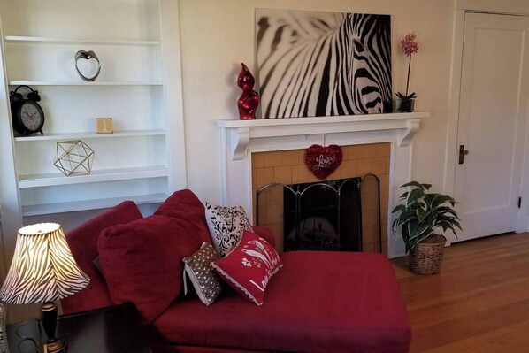 Una sala de estar acogedora con un sofá rojo, almohadones decorativos, un estampado de cebra en la pared, una decoración con forma de corazón sobre una chimenea y una planta en maceta.