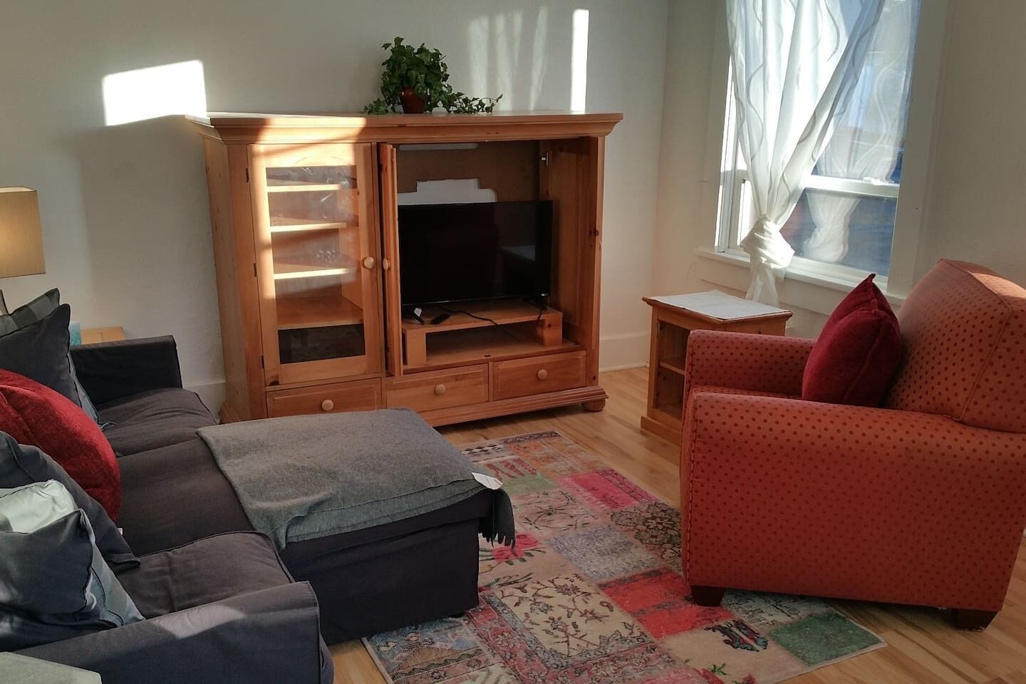 Una sala de estar con un mueble de madera para entretenimiento, un televisor, un sofá rojo estampado y un otoman gris.
