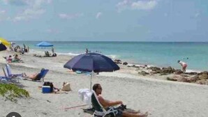 Ein Strandbild mit Menschen, die unter Sonnenschirmen entspannen, blauem Himmel und ruhiger See.