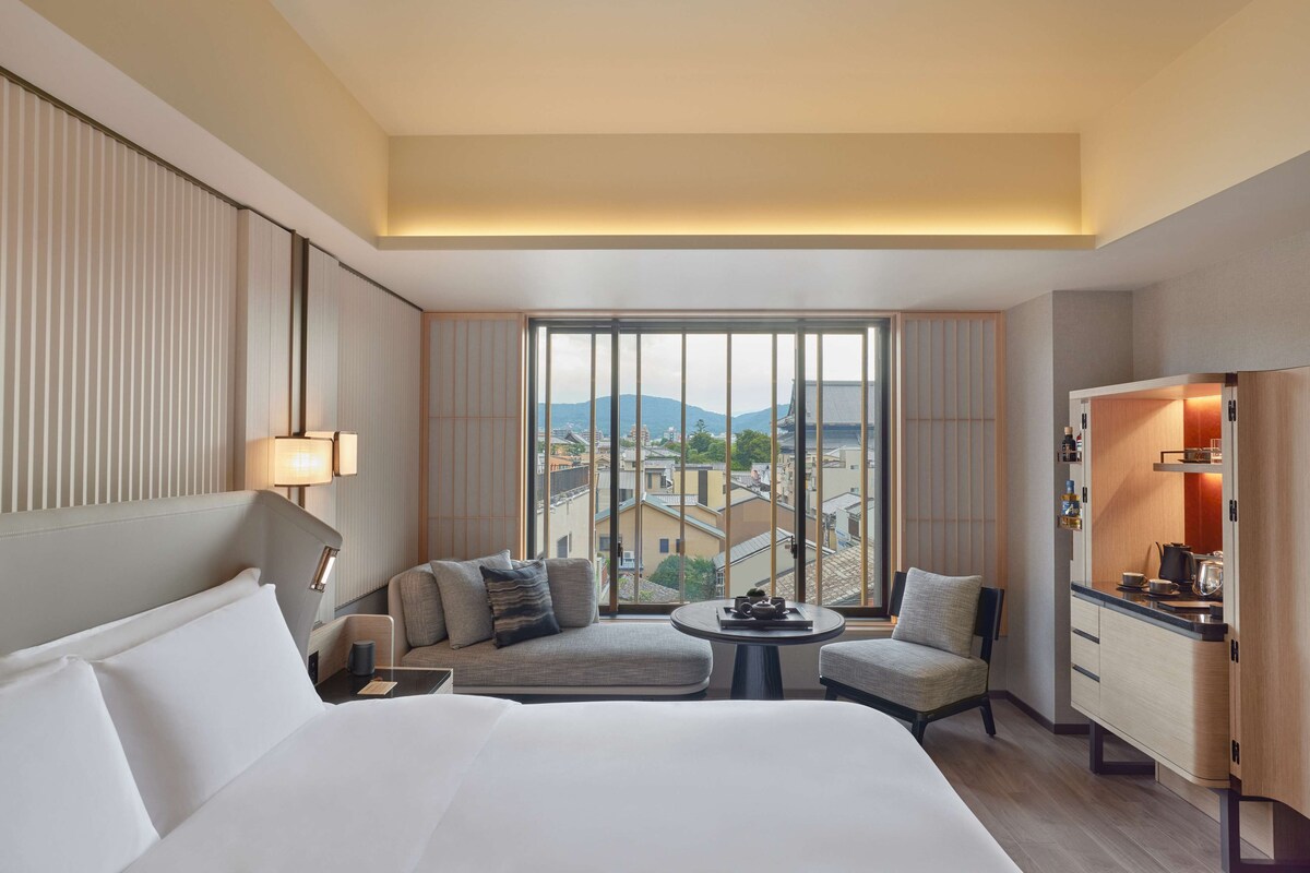 Une chambre d’hôtel comprenant un lit, un coin salon avec une table, un bureau avec une lampe et une vue sur les bâtiments et les montagnes.