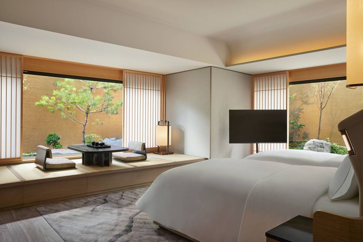 A modern hotel room with a large bed, a seating area with a table and chairs, a TV, and a view of trees through the windows.