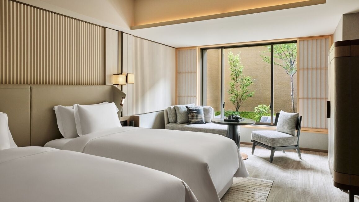 A hotel room with two beds, a seating area with a table, and large windows with a view of greenery.