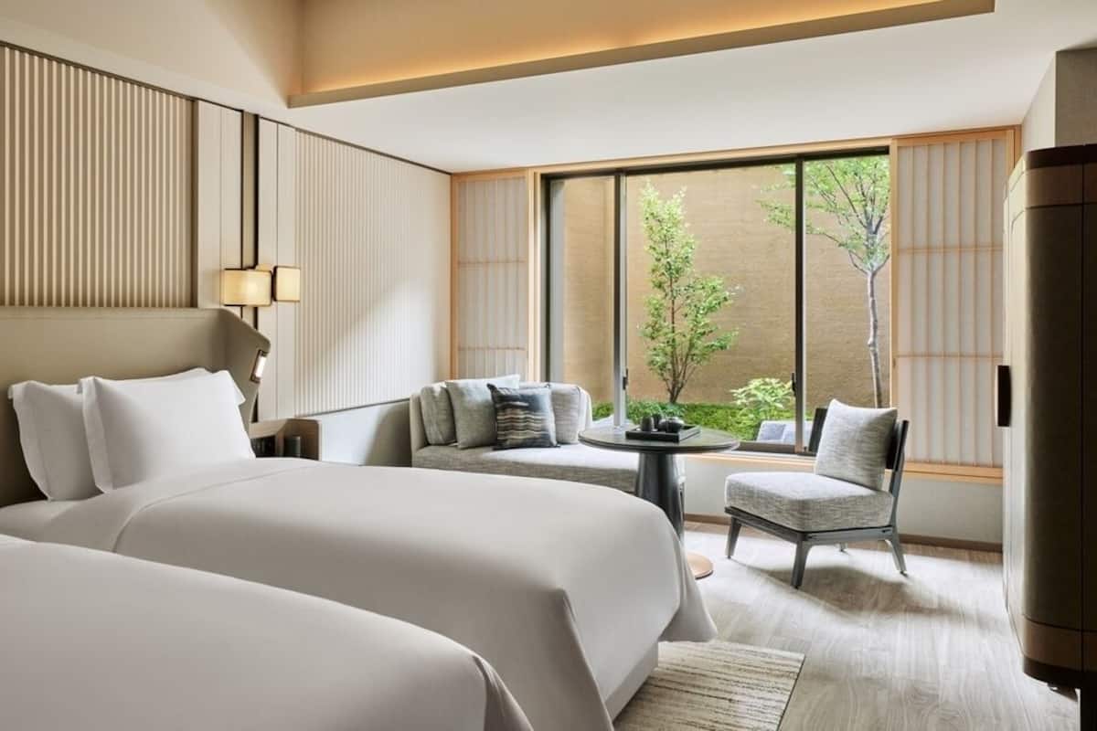 A hotel room with two beds, a seating area with a table, and large windows with a view of greenery.