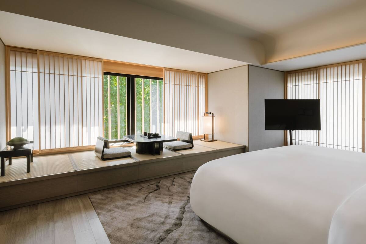 A modern hotel room with a large bed, a seating area with a table and chairs, a flat-screen TV, and large windows with vertical blinds.
