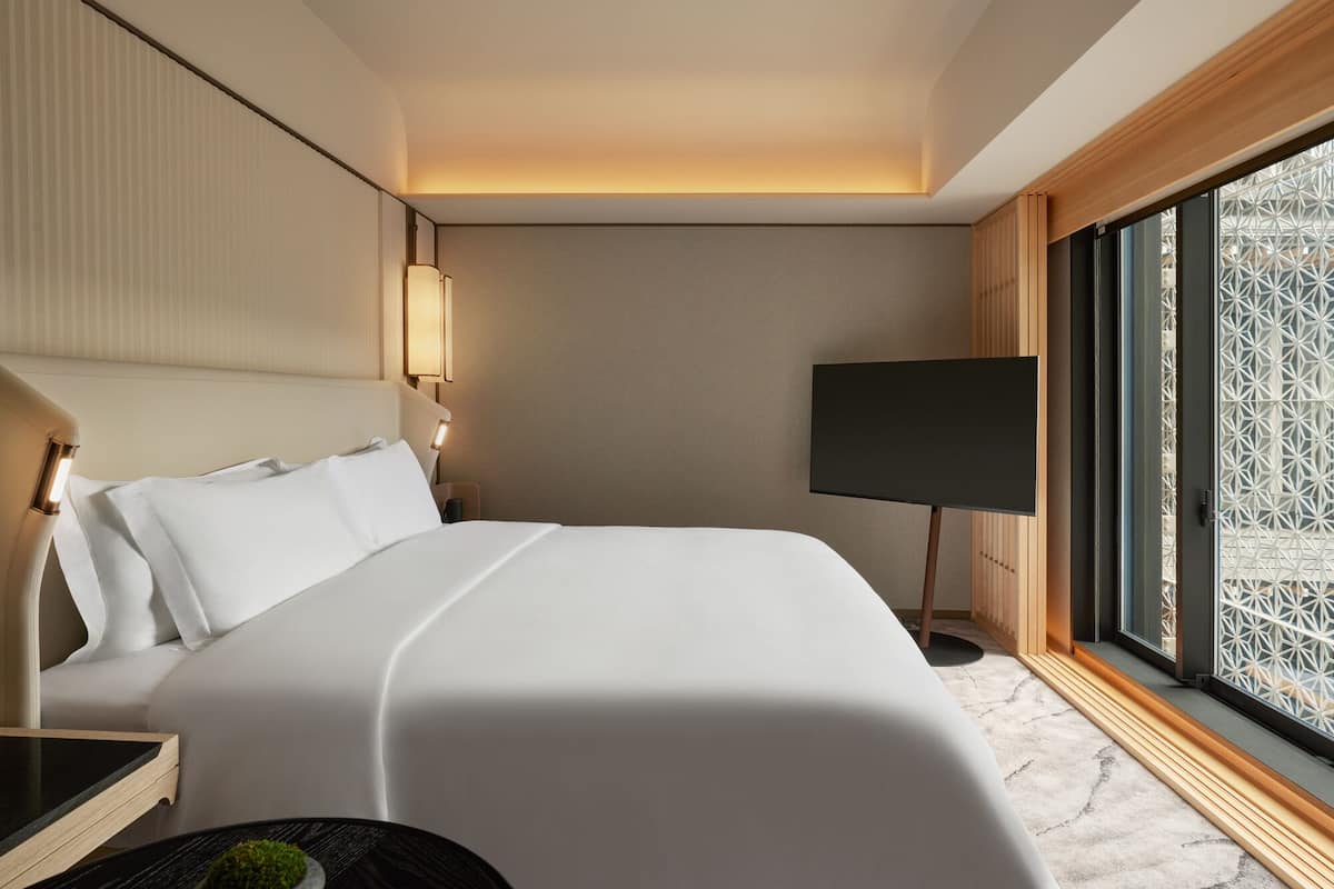 A modern hotel room with a large bed, a bedside table, a TV, and a window with a view of a geometric structure outside.