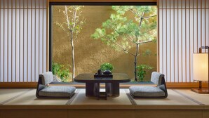 Um quarto japonês tradicional com piso de tatame, uma mesa baixa, duas cadeiras e uma janela grande com vista para o verde.
