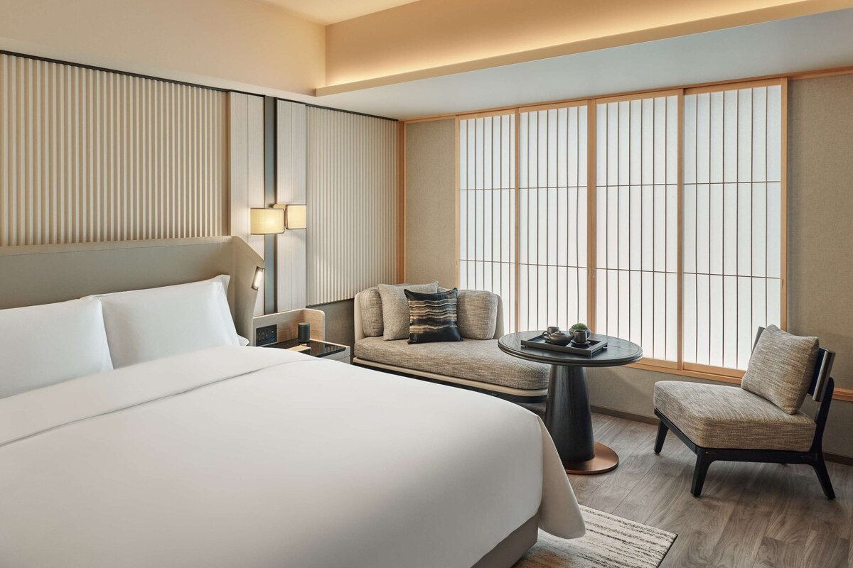 Une chambre d’hôtel avec un grand lit, un canapé, une chaise, une petite table et une fenêtre avec des stores verticaux.