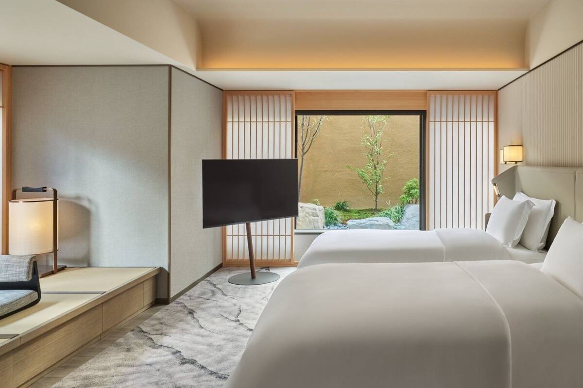 A hotel room with a bed, a TV, a desk, and a view of greenery outside.