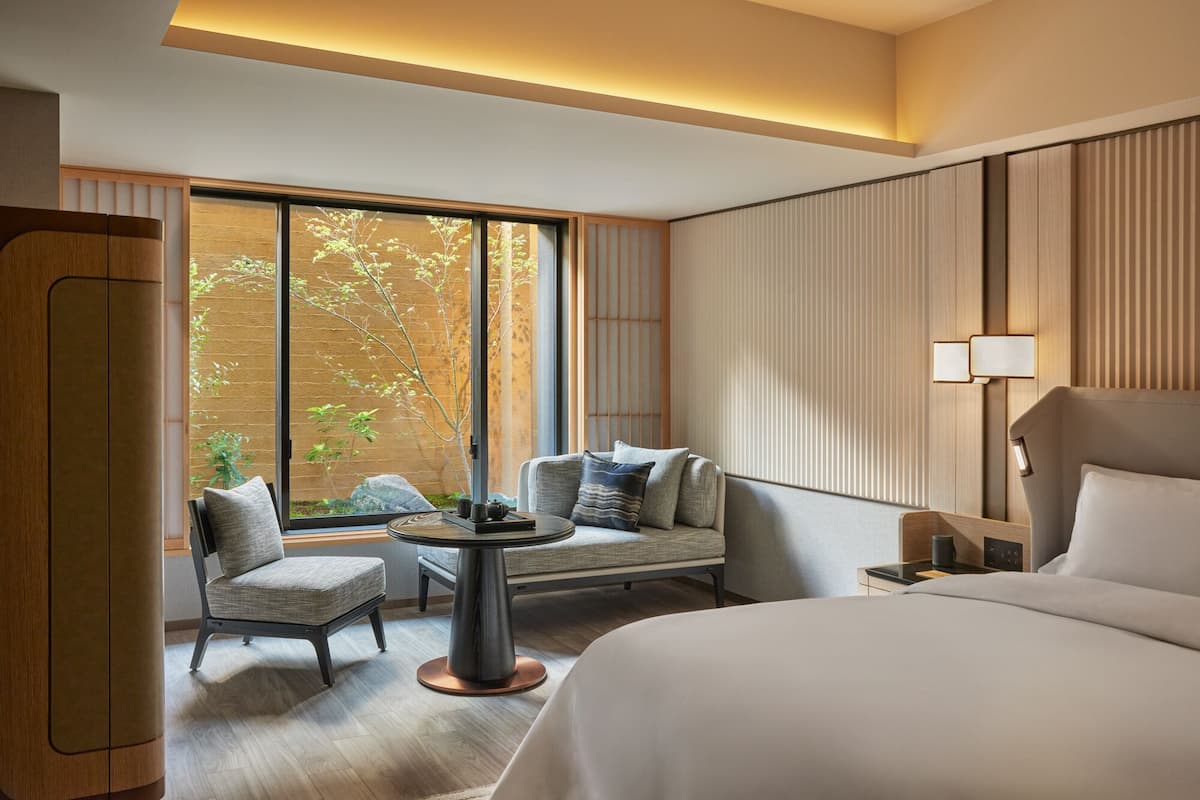 A hotel room with a large bed, a seating area with a table, and a view of greenery through a large window.