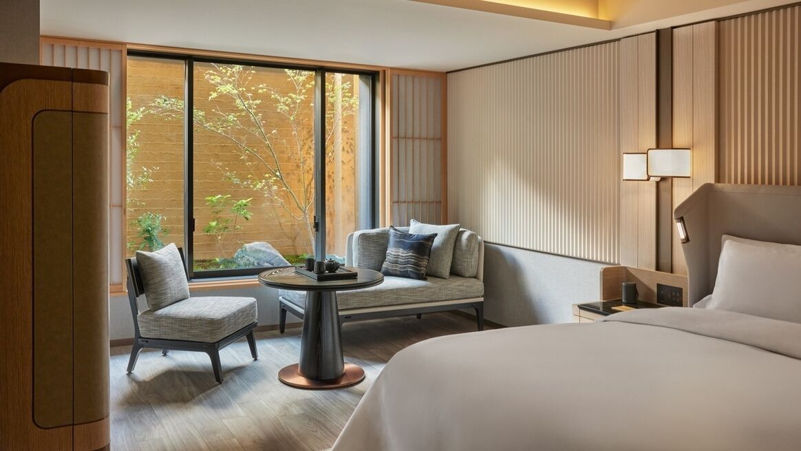 A hotel room with a bed, a sofa, a small table, and a window with a view of trees.