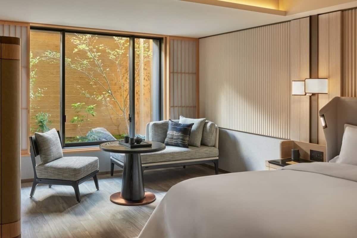 A hotel room with a bed, a sofa, a small table, and a window with a view of trees.