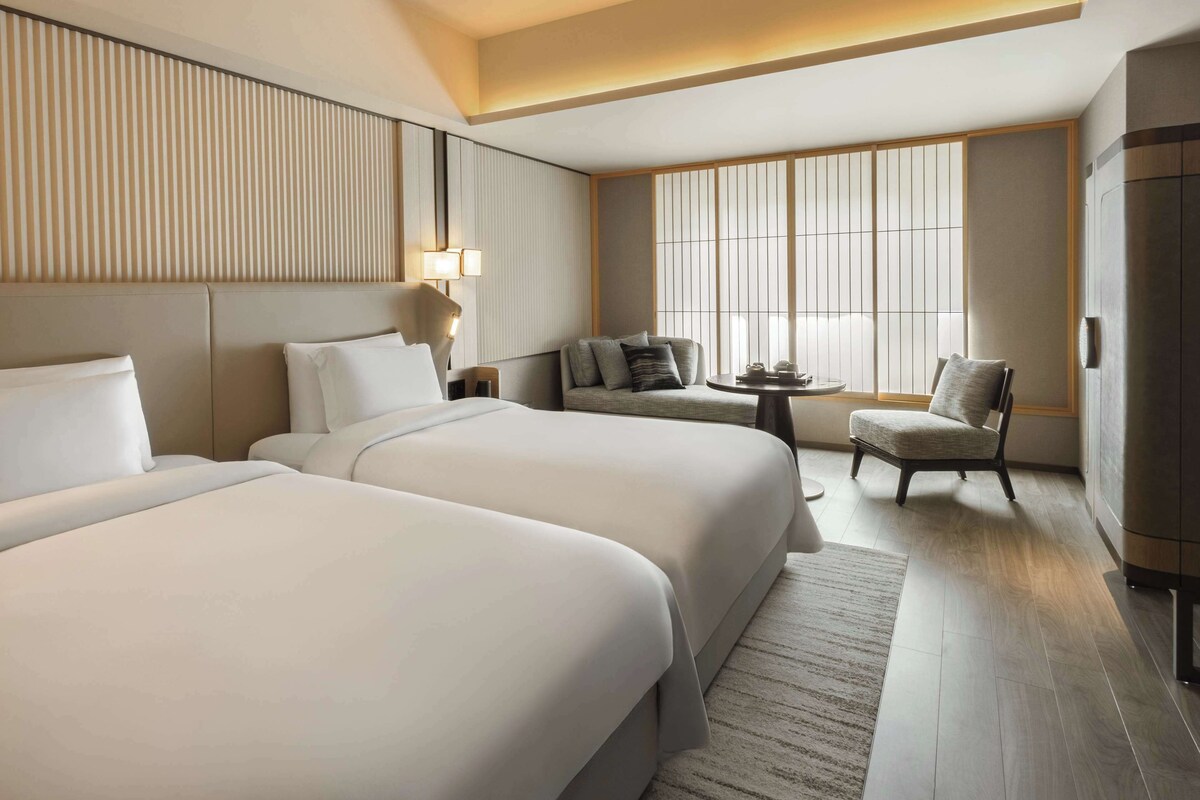 Une chambre d’hôtel avec deux lits, un canapé, une table à manger et une vue dégagée sur l’extérieur grâce à de grandes fenêtres.