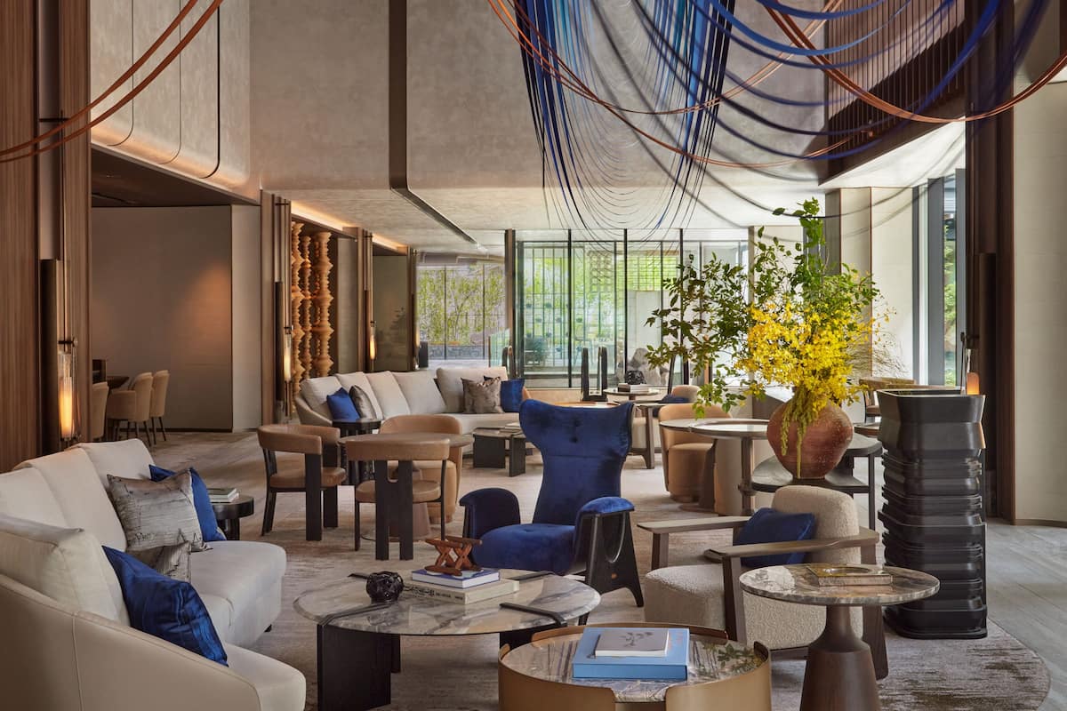 A modern lounge area with sofas, armchairs, and tables, featuring a large window with a view of greenery.