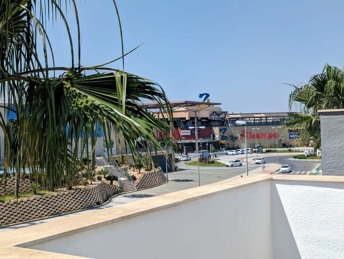 Une vue d’un parking avec un bâtiment en arrière-plan, un palmier et un ciel dégagé.
