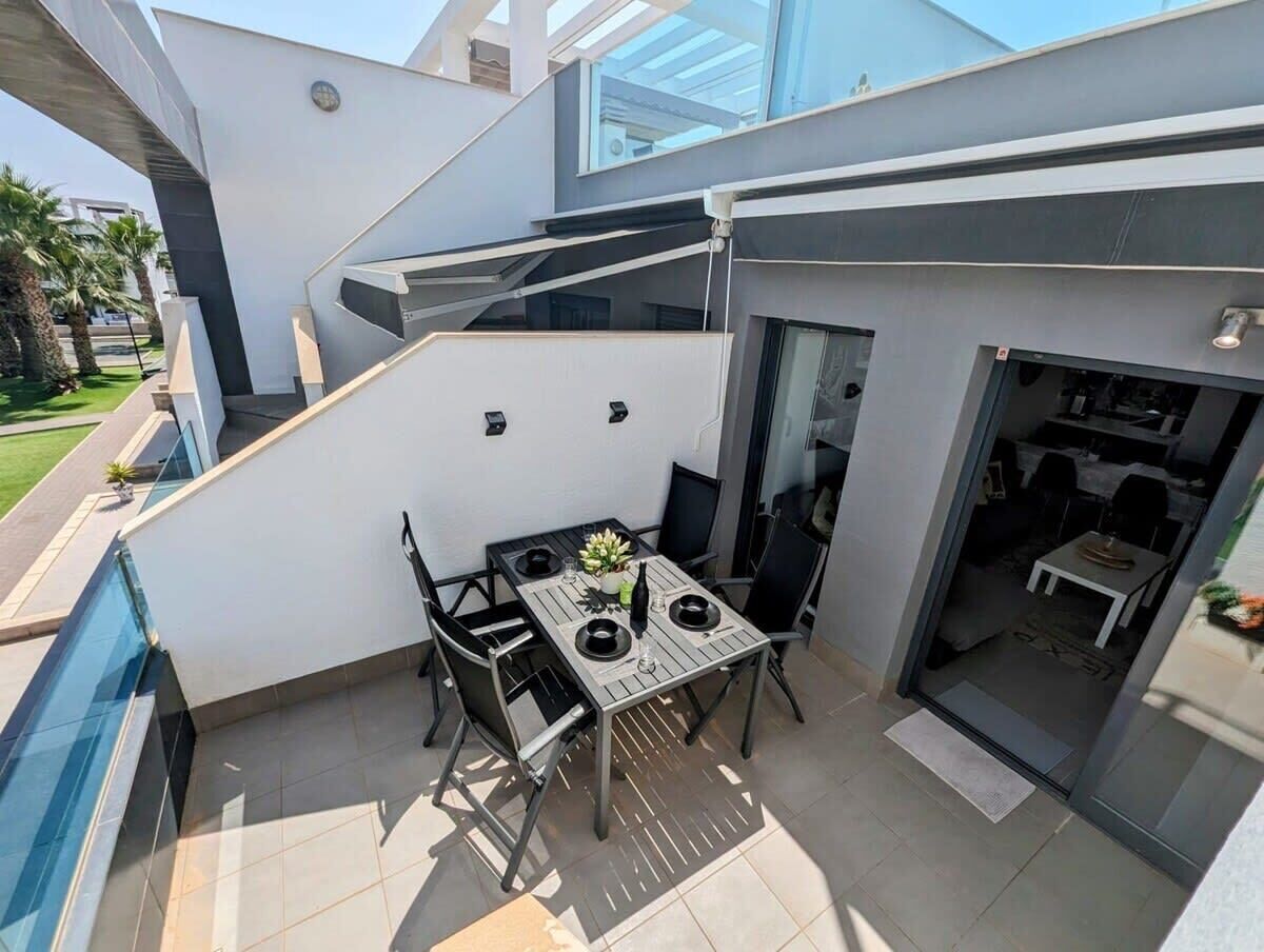 A modern outdoor patio with a dining table and chairs, a small garden area, and a glass-enclosed space leading to an interior room.