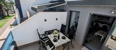 A modern outdoor patio with a dining table and chairs, a small garden area, and a glass-enclosed space leading to an interior room.