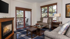 Ein Wohnzimmer mit Kamin, einer Couch, einem Couchtisch und einem Fernseher. Es gibt einen Balkon mit einem Sessel und einem Tisch.