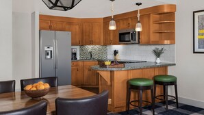 Eine moderne Küche mit Holzausbau, einem Edelstahlkühlschrank, einer Mikrowelle und einer Arbeitsplatte aus Granit. Es steht ein Esstisch mit Stühlen und eine Schüssel mit Obst bereit.