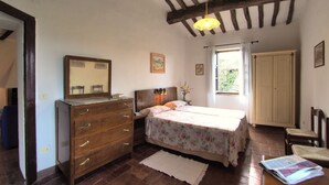 A bedroom with a wooden dresser, a bed with floral bedding, a mirror, and a window with curtains. - Castello di Montalto - Casa di Giocche (Castelnuovo Berardenga)