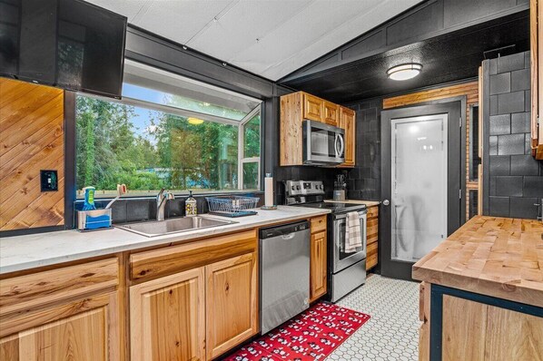 A modern kitchen with wooden cabinets, stainless steel appliances, and a large window offering a view of greenery.