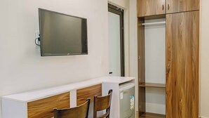 Een kleine kamer met een bureau, twee stoelen, een aan de muur bevestigde televisie en een houten garderobe.