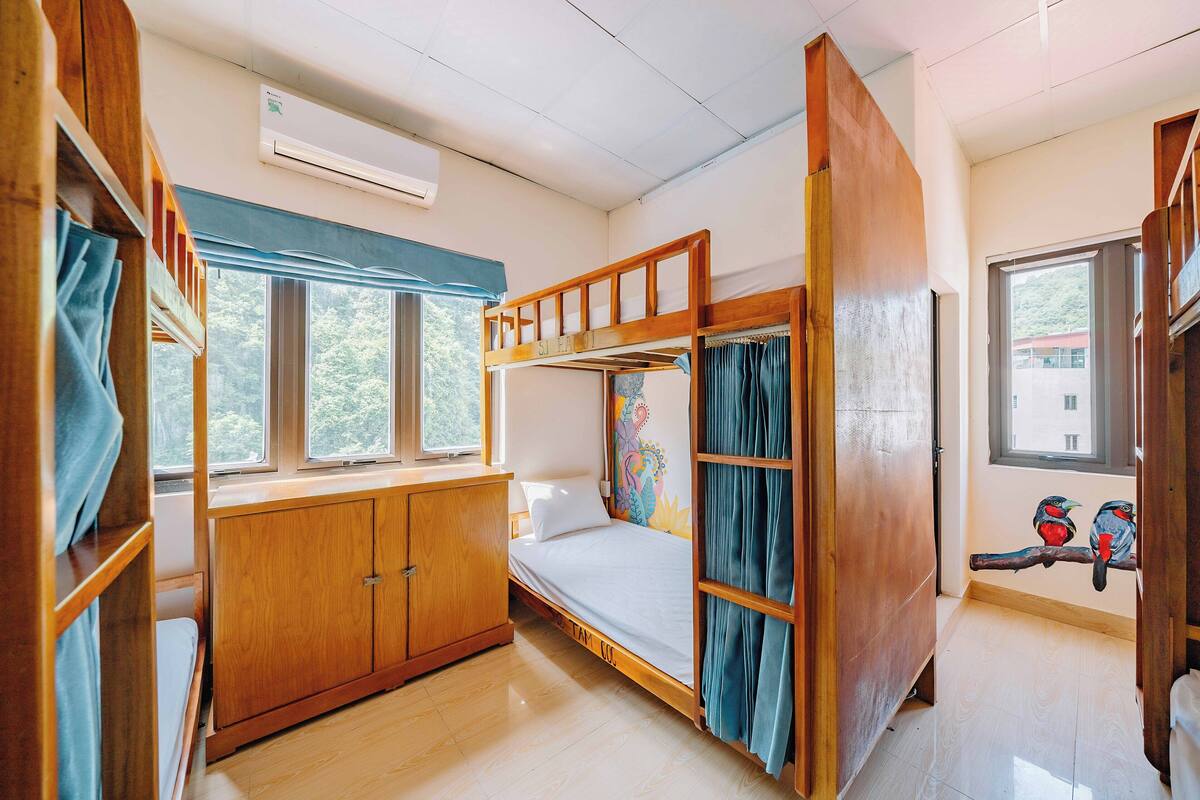 Een slaapkamer met een stapelbed, een houten garderobe, een raam met gordijnen en een wandairconditioner.