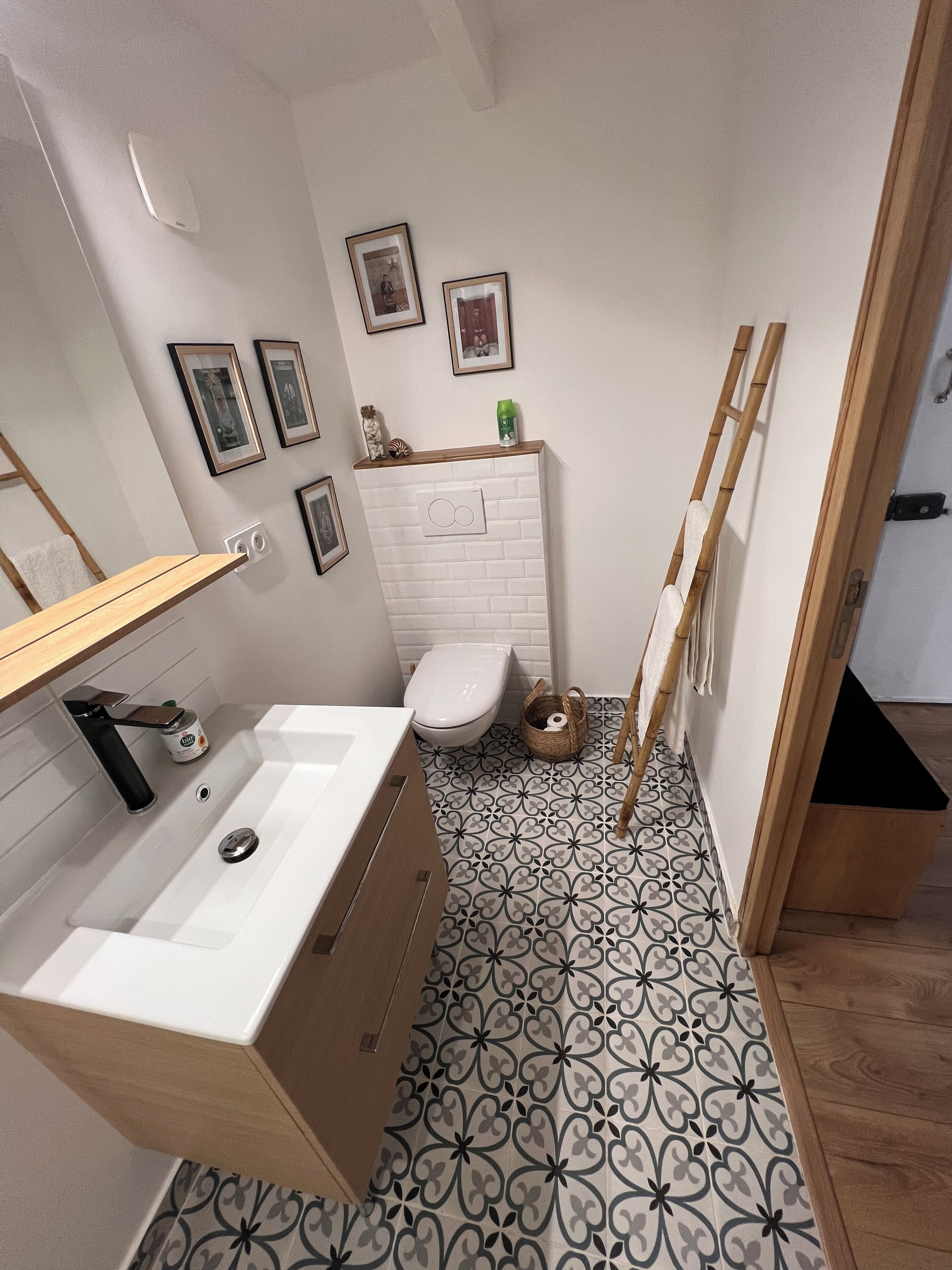 Et lille badeværelse med en hvid vask, toilet og spejl. Der er billeder i rammer på væggen, og en træstige står lænet op ad væggen.