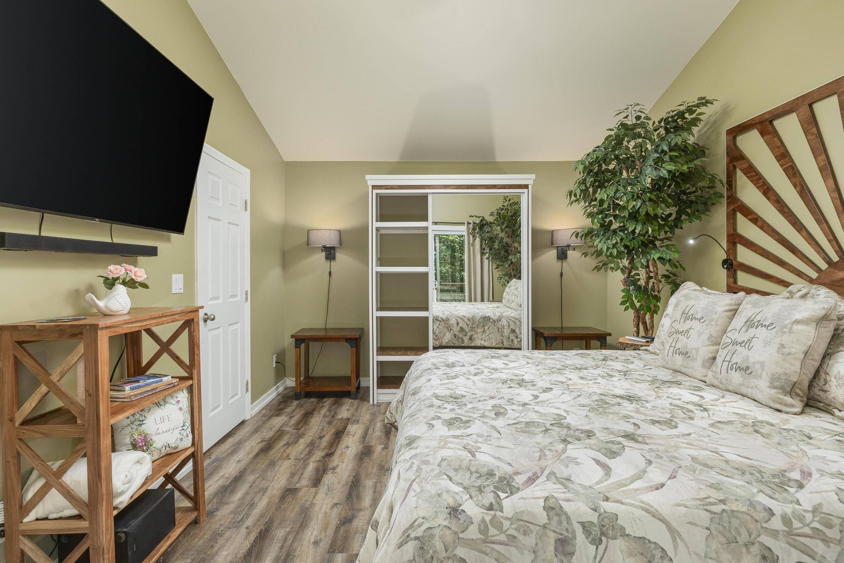 A bedroom with a bed, a TV, a wooden side table, a potted plant, and a sliding door leading to a balcony.