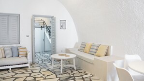 Ein Zimmer mit weißen Wänden und einem Steinboden, ein weißes Sofa mit gestreiften Kissen, ein kleiner weißer Tisch und eine Treppe, die zu einer anderen Ebene führt.