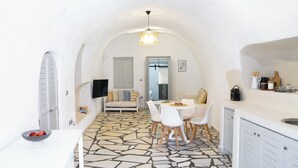 Eine kompakte Küche mit Steinplattenboden, ein Essbereich mit weißen Stühlen, eine Couch und ein an der Wand befestigter Fernseher.