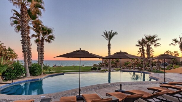 A pool area with lounge chairs and umbrellas, surrounded by palm trees and a view of the ocean.