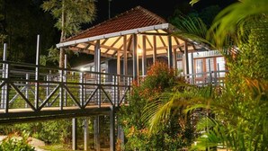 A covered outdoor area with a wooden deck and a metal railing, surrounded by lush greenery. - B'camp Resorts & Homestays (Vythiri)