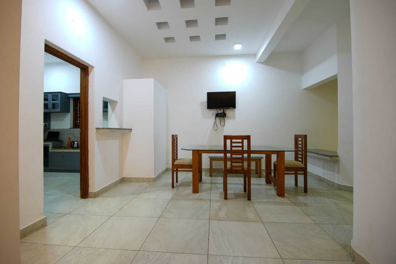 A dining area with a table and chairs, a TV mounted on the wall, and a kitchen visible through an open doorway.