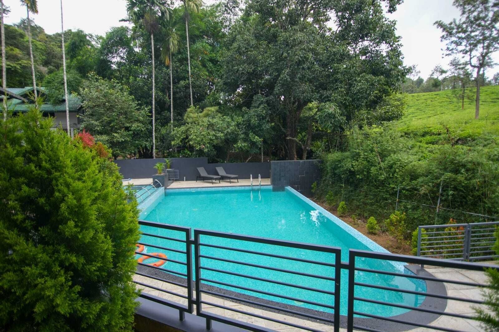 Una piscina rectangular con agua azul cristalina, rodeada por una cerca de metal negro. Hay dos sillas de descanso y un pequeño puesto de salvavidas cerca de la piscina.