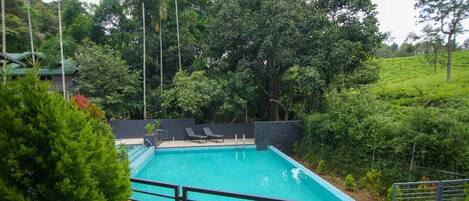 Una piscina rectangular con agua azul cristalina, rodeada por una cerca de metal negro. Hay dos sillas de descanso y un pequeño puesto de salvavidas cerca de la piscina.
