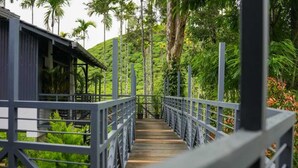 A wooden bridge with metal railings leading to a building surrounded by lush greenery and palm trees. - B'camp Resorts & Homestays (Vythiri)