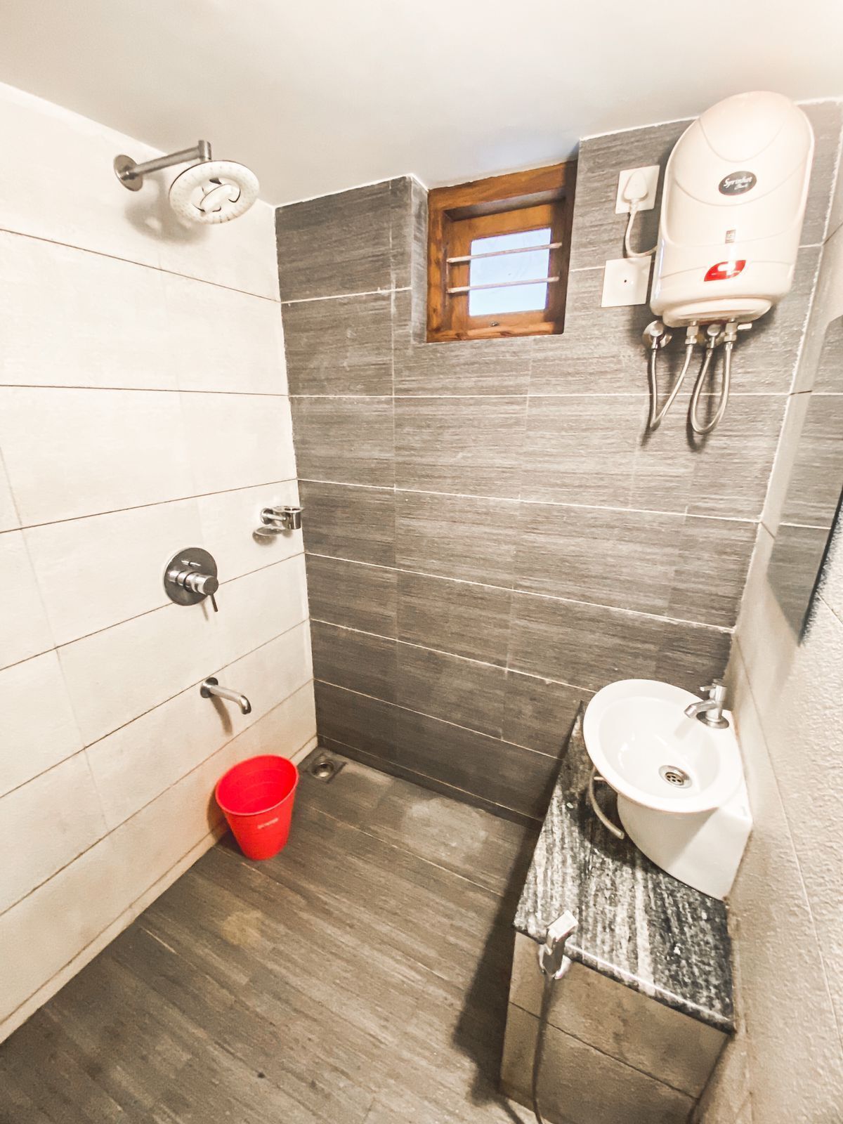 Une petite salle de bain avec un lavabo blanc, un seau rouge, une pomme de douche et une fenêtre.