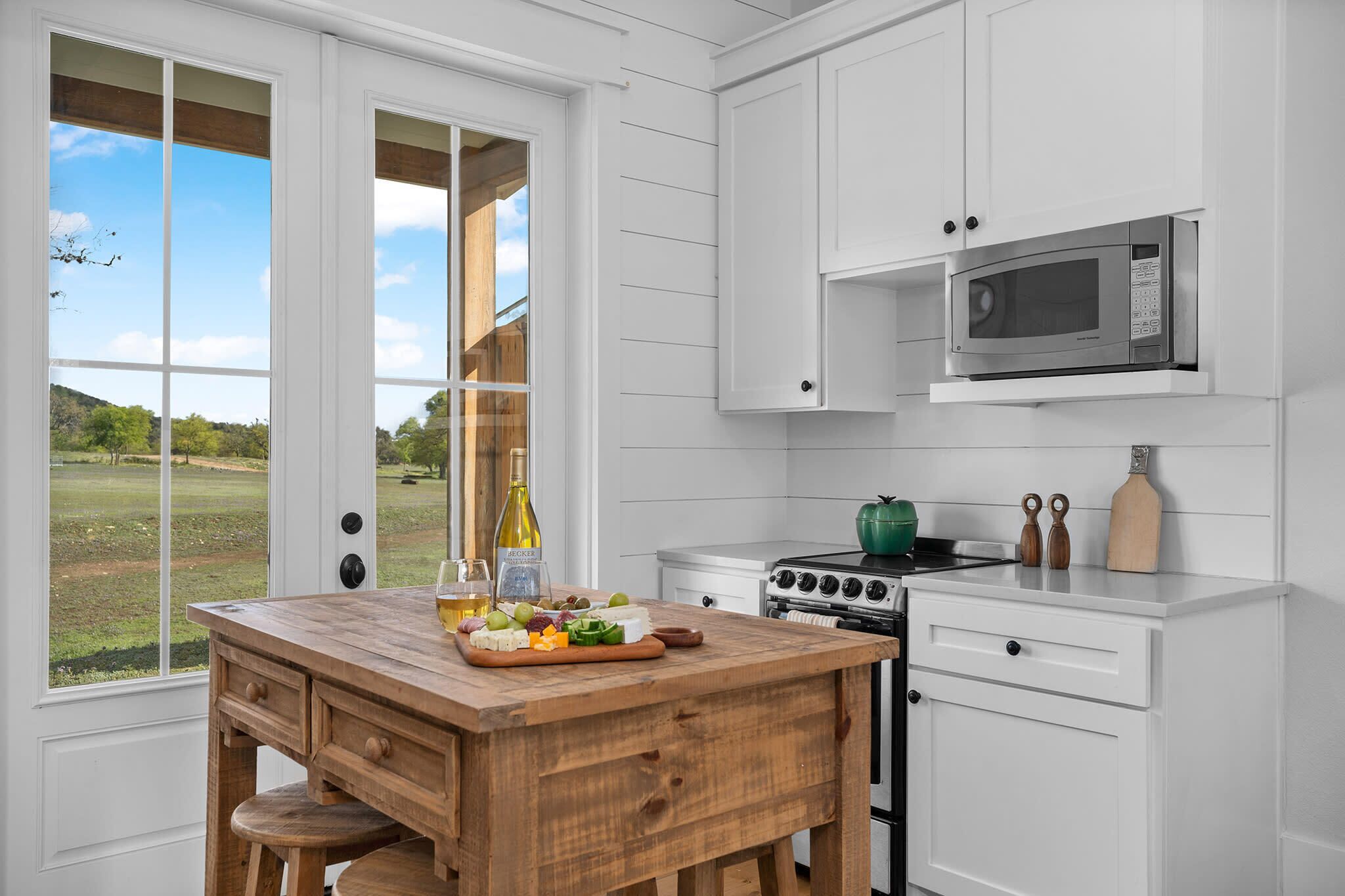 A kitchen with white cabinets, a wooden island with a fruit platter, a microwave, a stove, and a view of a grassy field through large windows.