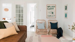 A cozy living room with a brown sofa, two wicker chairs, a pink table, and a poster on the wall advertising a wine and cheese club.