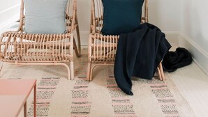 A room with two wicker chairs, one with a light blue pillow and the other with a dark blue pillow. A black blanket is draped over the back of one chair. A framed poster on the wall reads "WINE & CHEESE CLUB" with a wine bottle and cheese illustration, and "WEEKNIGHTS 7:30-11:00 (SOMETIMES WEEKENDS)" text.