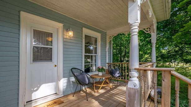 A porch with a wooden deck, two chairs, a small table, and a white door.