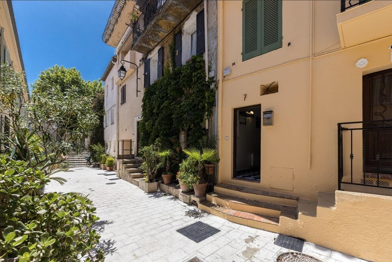 A narrow street with buildings on either side, potted plants, and a clear blue sky.