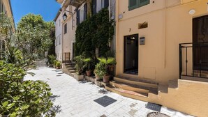 A narrow street with buildings on either side, potted plants, and a clear blue sky.