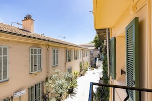 A narrow street with buildings on either side, featuring balconies with potted plants and shutters.
