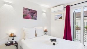 A hotel room with a bed, bedside tables, a painting, an air conditioning unit, and a view of the outside.